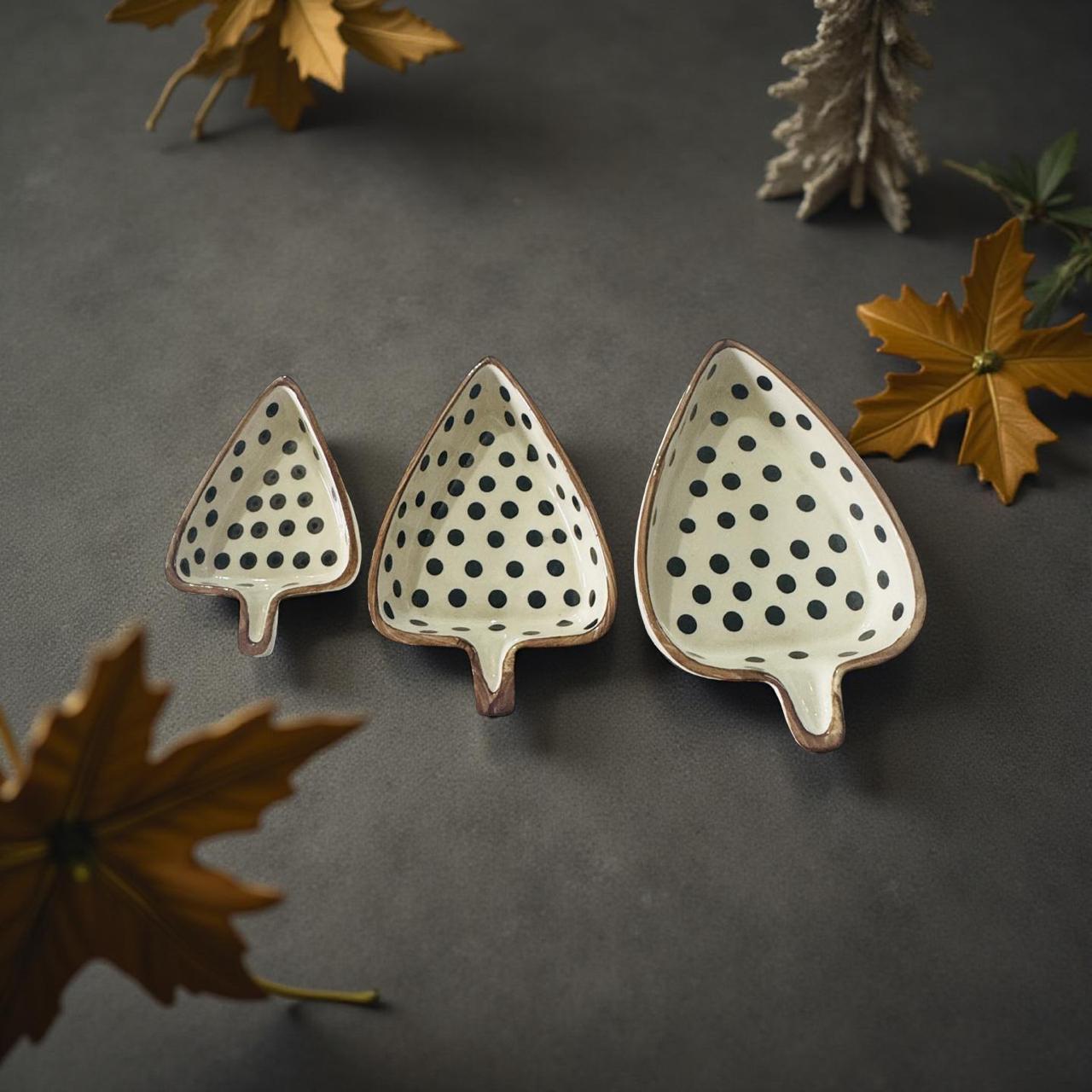 Three leaf-shaped ceramic dishes with black polka dots on a dark surface with autumn leaves.
