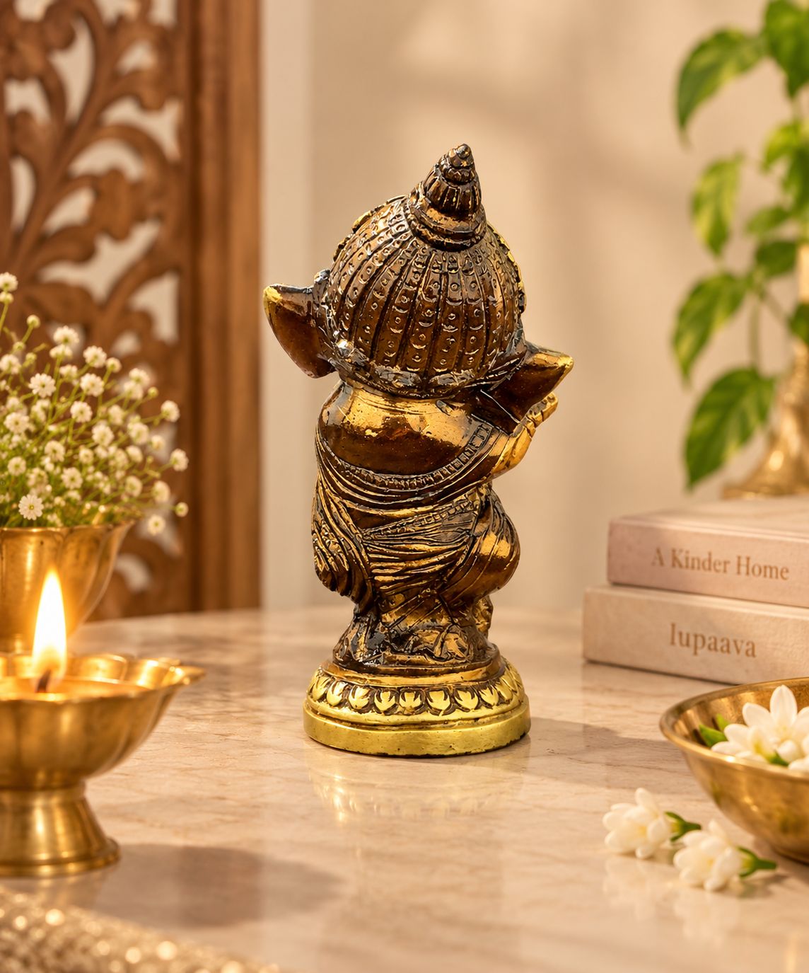 A small metallic Ganesha figurine with a gold and bronze finish, standing on a marble surface with decorative props including a lit candle, flowers, and books in the background.