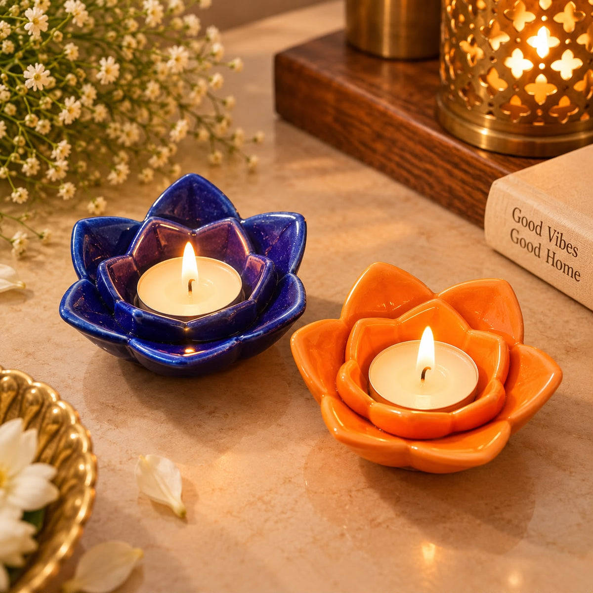 Two ceramic lotus-shaped tea light candle holders, one in glossy cobalt blue and one in glossy terracotta orange, each holding a lit tealight candle on a warm beige tabletop with decorative flowers and a lantern in the background.
