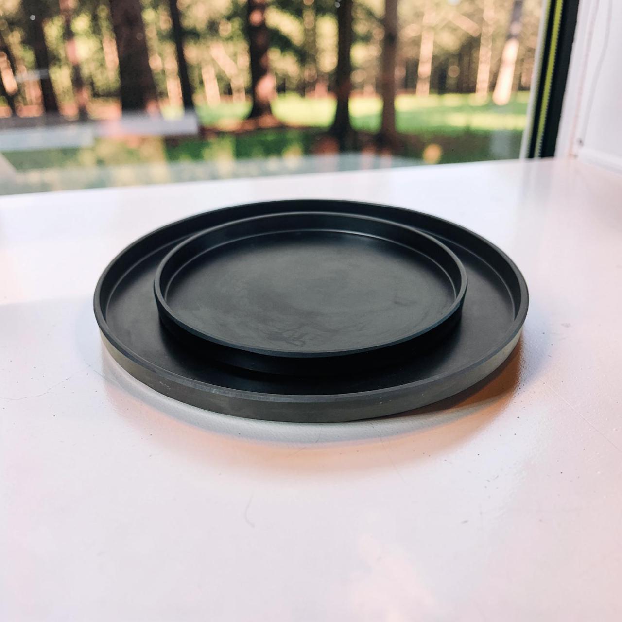 Two black ceramic trays stacked on a white surface with a blurred outdoor background