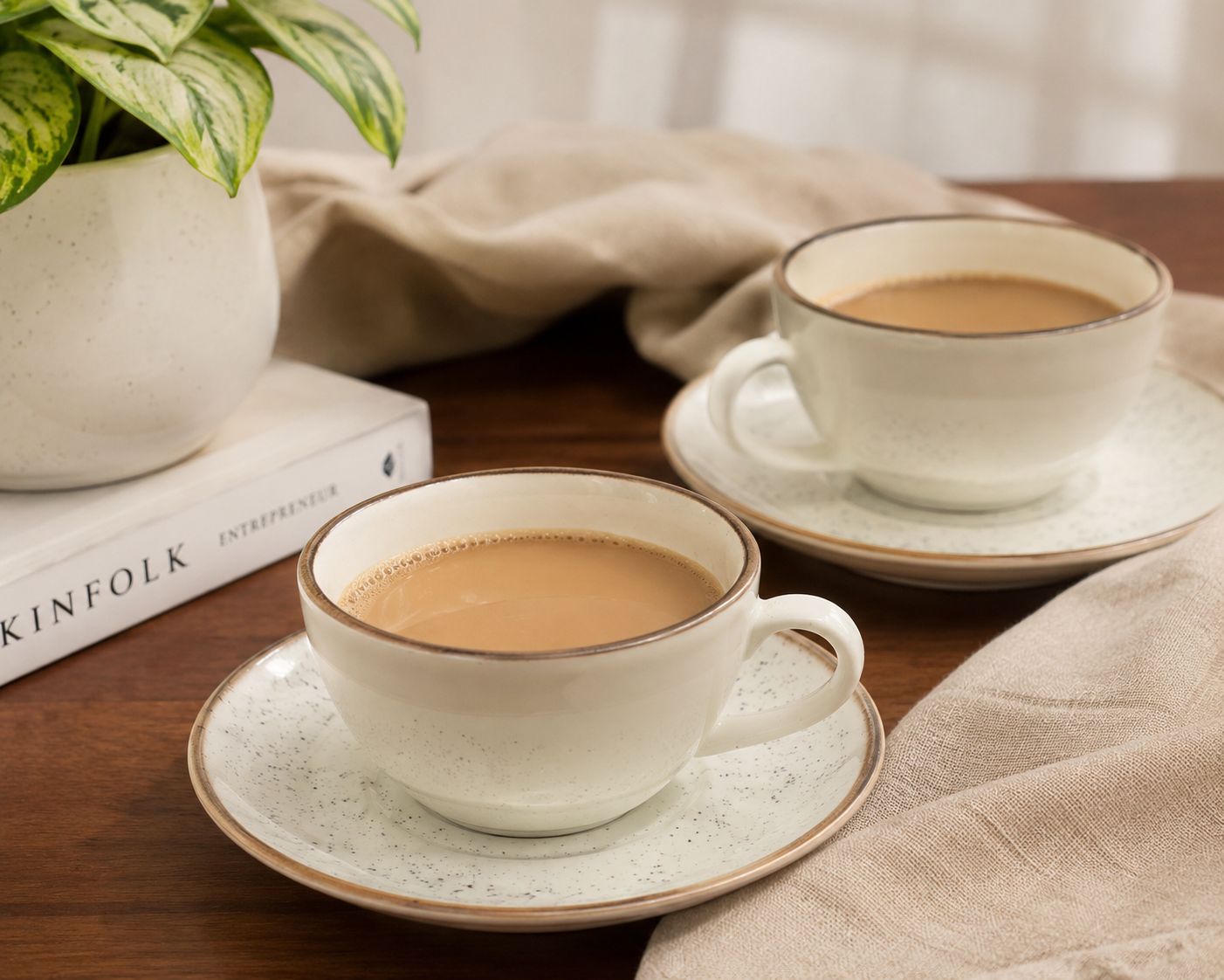 Two cups of coffee on saucers on a wooden table with a plant and books in the background.