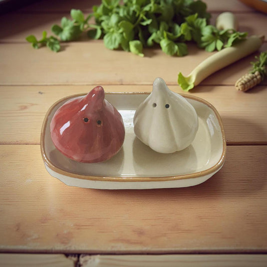 Two ceramic garlic-shaped salt and pepper shakers on a wooden surface with greenery in the background.