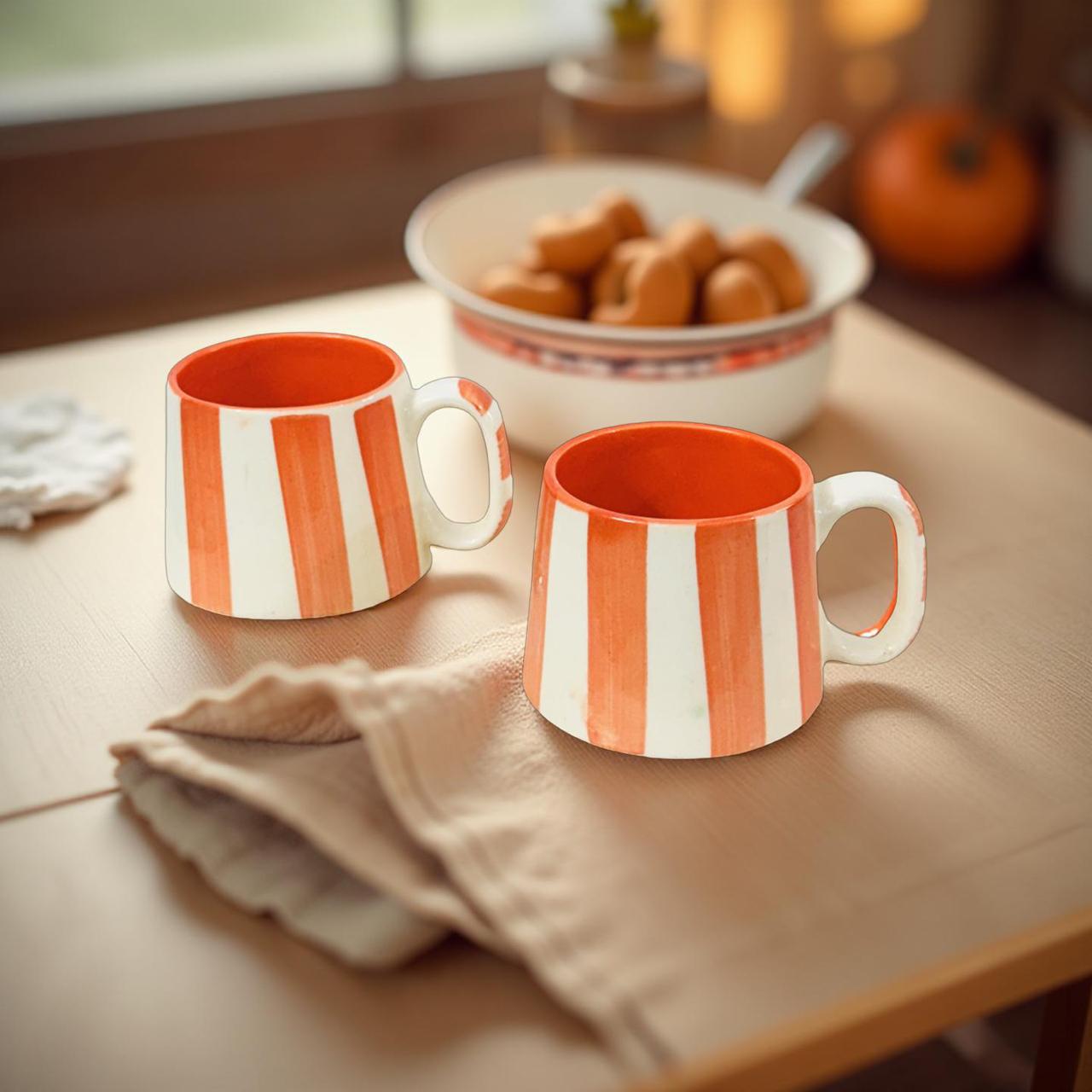 Two orange and white striped mugs on a wooden table with a bowl of pastries in the background.