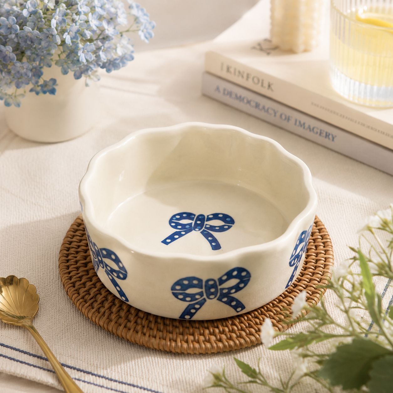 A white ceramic bowl with a scalloped rim and blue bow patterns, displayed on a woven round mat with a gold spoon, flowers, and books in the background.