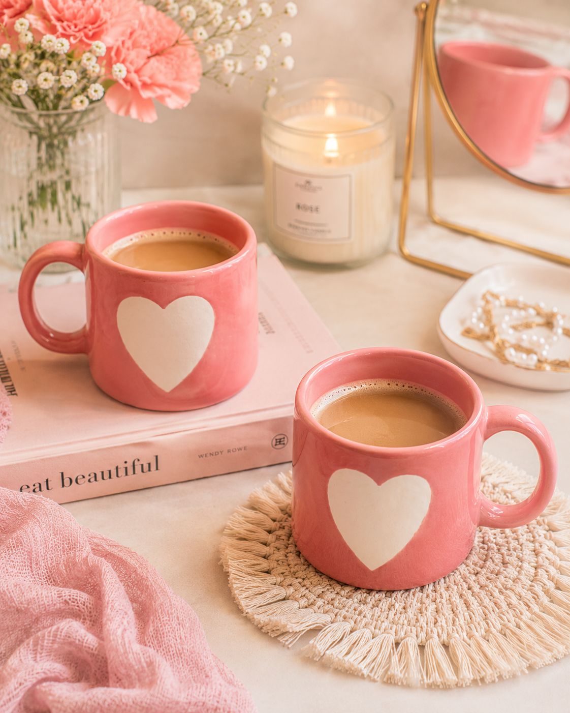 Two pink ceramic mugs filled with coffee, each featuring a large white heart on the front, styled on a table with a book, candle, flowers, and a mirror in a warm, cozy setting.