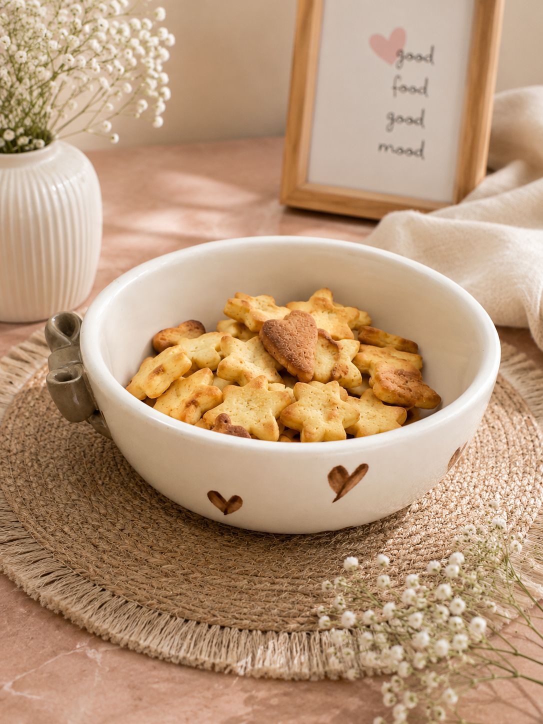 A white ceramic bowl with heart-shaped cutouts and a small metal handle, filled with star-shaped cookies, styled on a woven placemat with baby's breath flowers and a framed sign in the background.
