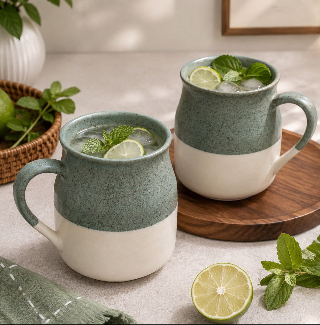 Two ceramic mugs with a two-tone sage green and cream finish, filled with iced drinks and garnished with lime slices and mint leaves, styled on a tabletop with a wooden board, a folded green cloth, and fresh mint.