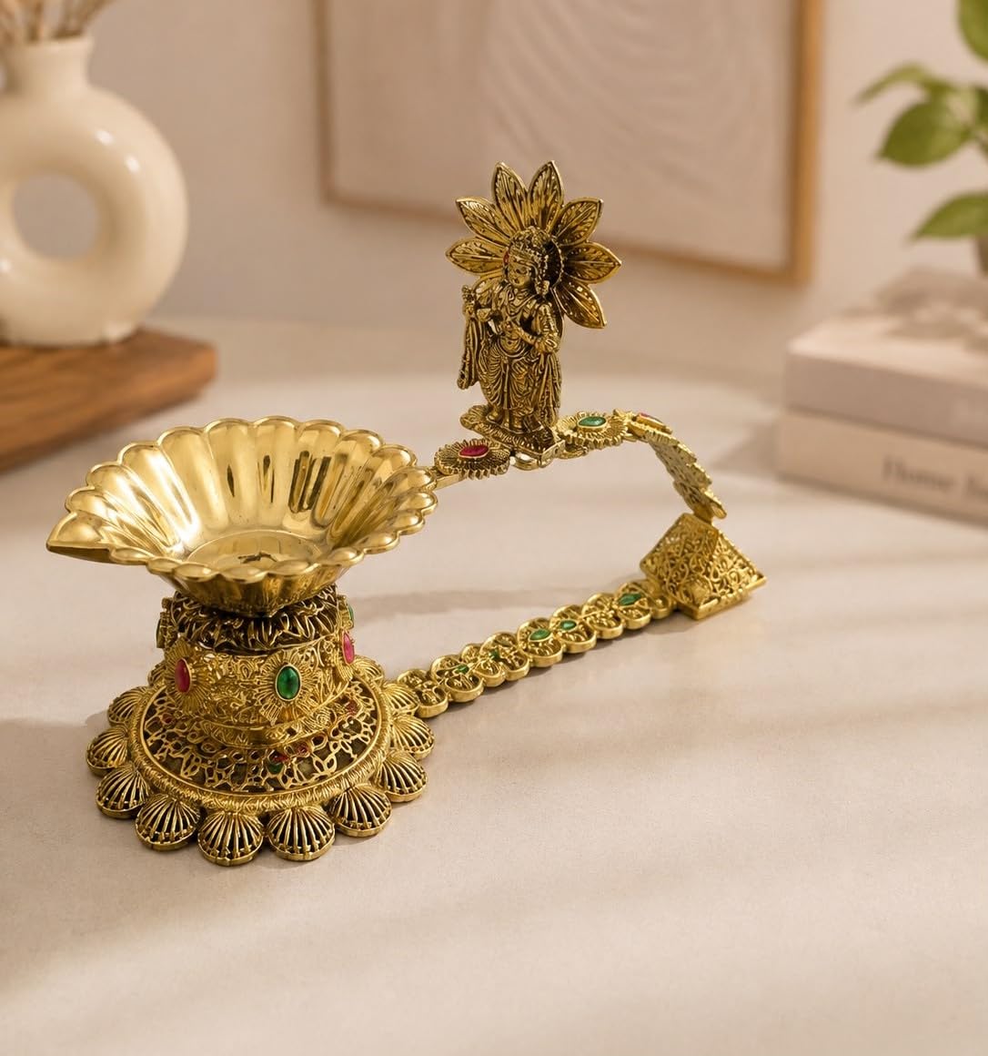 An ornate brass oil lamp holder with a decorative floral finial and a chain connecting the bowl to the stand, accented with small green and red stones, displayed on a light beige surface.