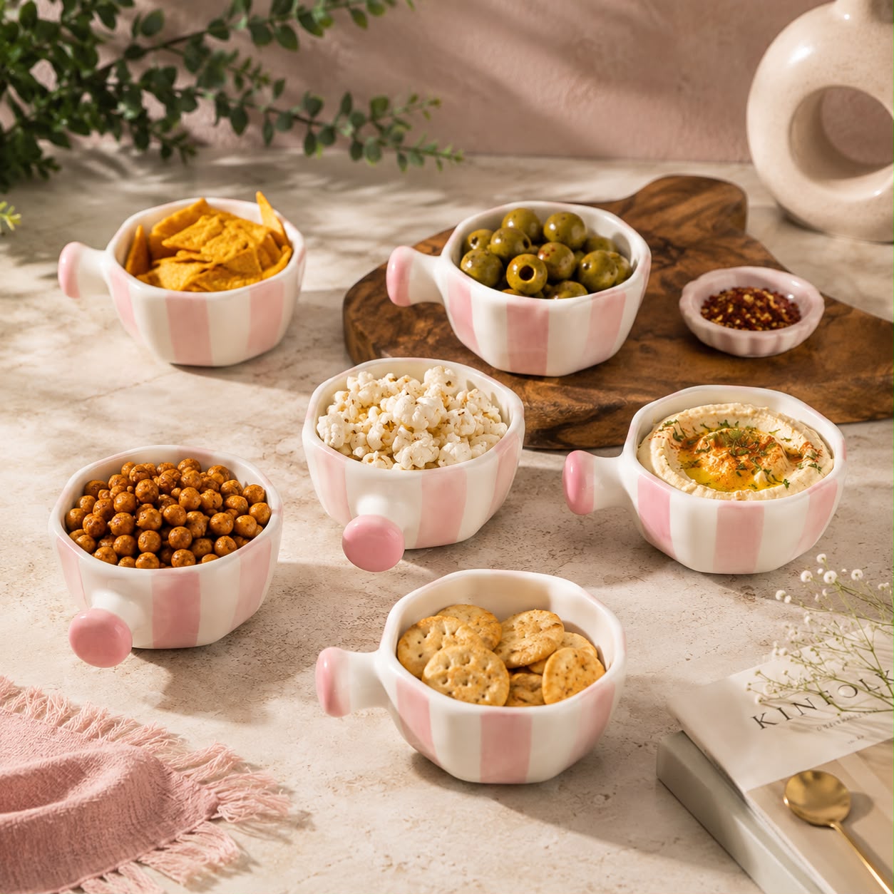 Six small ceramic bowls with pink and white stripes are arranged on a tabletop, filled with snacks such as crackers, popcorn, olives, chickpeas, and dips. The scene includes a wooden board, a napkin, a spoon, and soft decorative props in a styled indoor setting.
