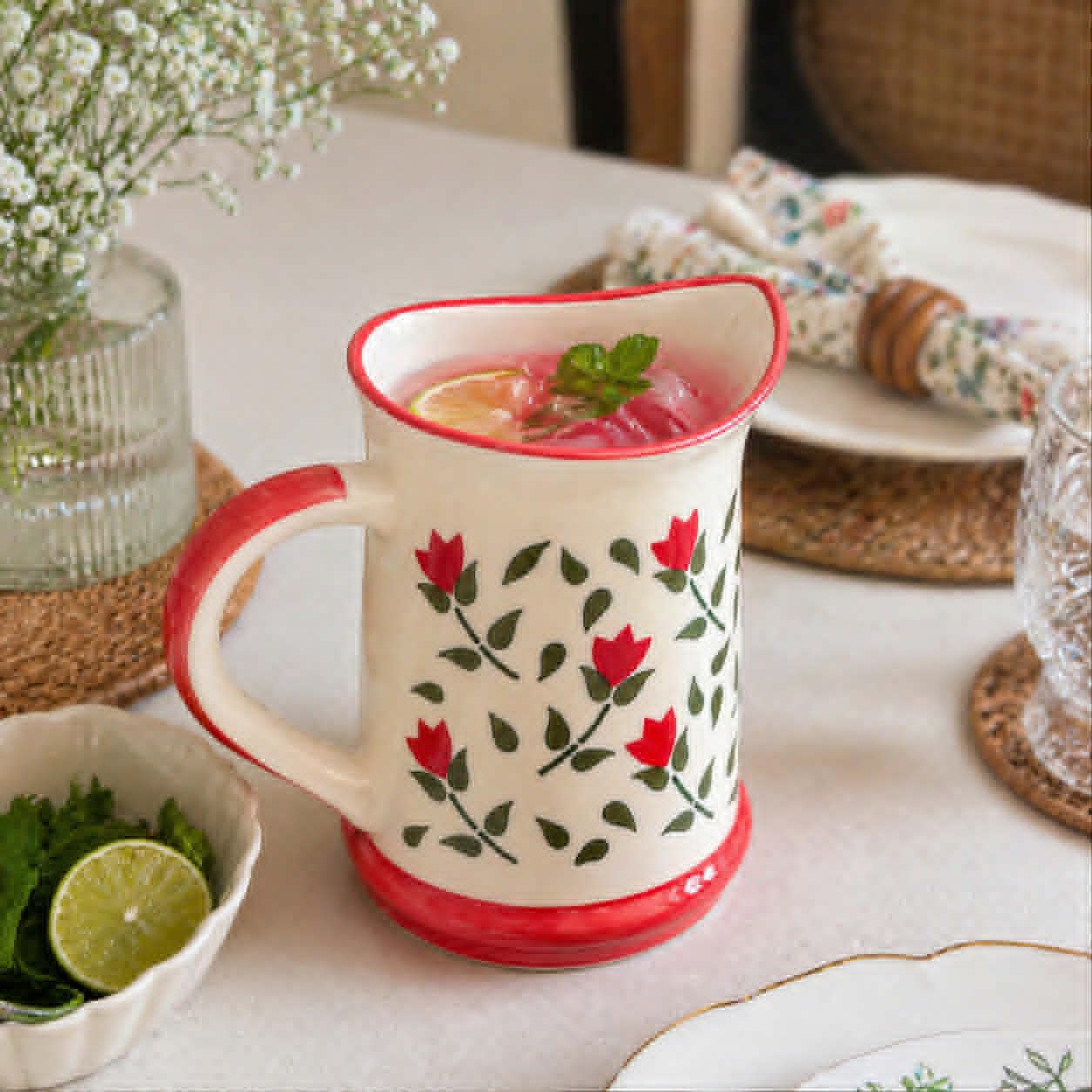 Decorative mug with floral pattern on a table with a bowl of limes and a vase of flowers.