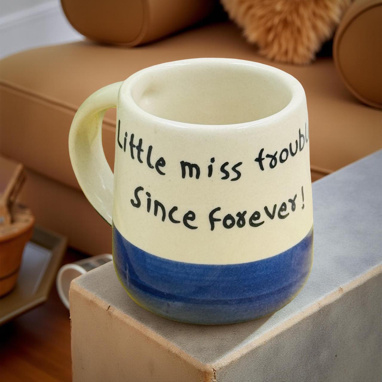 Ceramic mug with text 'Little miss trouble Since forever!' on a wooden block.