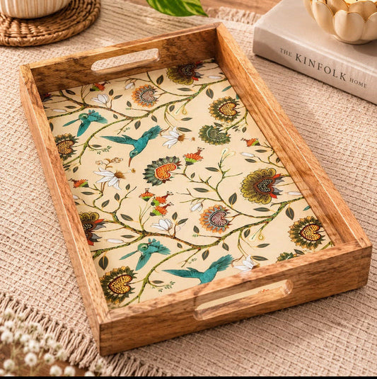 A rectangular wooden serving tray with cutout handles, featuring a cream-colored printed surface decorated with a botanical and bird pattern in blue, green, orange, and brown. The tray is styled on a woven beige table runner next to a potted green plant, a book, and a lit candle.