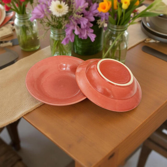 Two pink ceramic dishes on a wooden table with floral arrangements in the background.