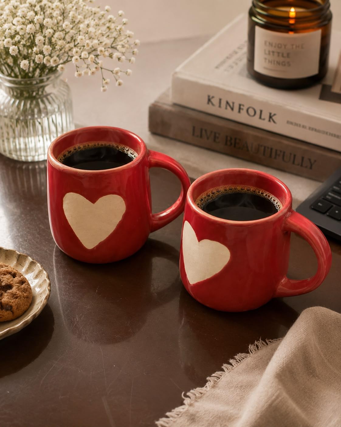 Two red ceramic mugs filled with black coffee sit on a dark tabletop in a cozy styled setting. Each mug has a large cream heart on the front.