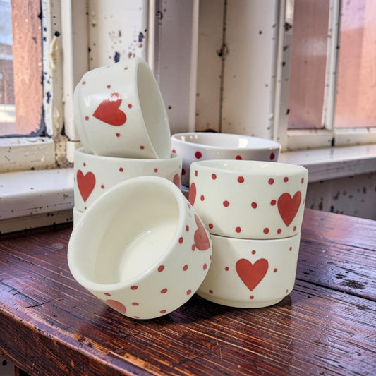 Set of ceramic bowls with red heart designs on a wooden surface near a window.