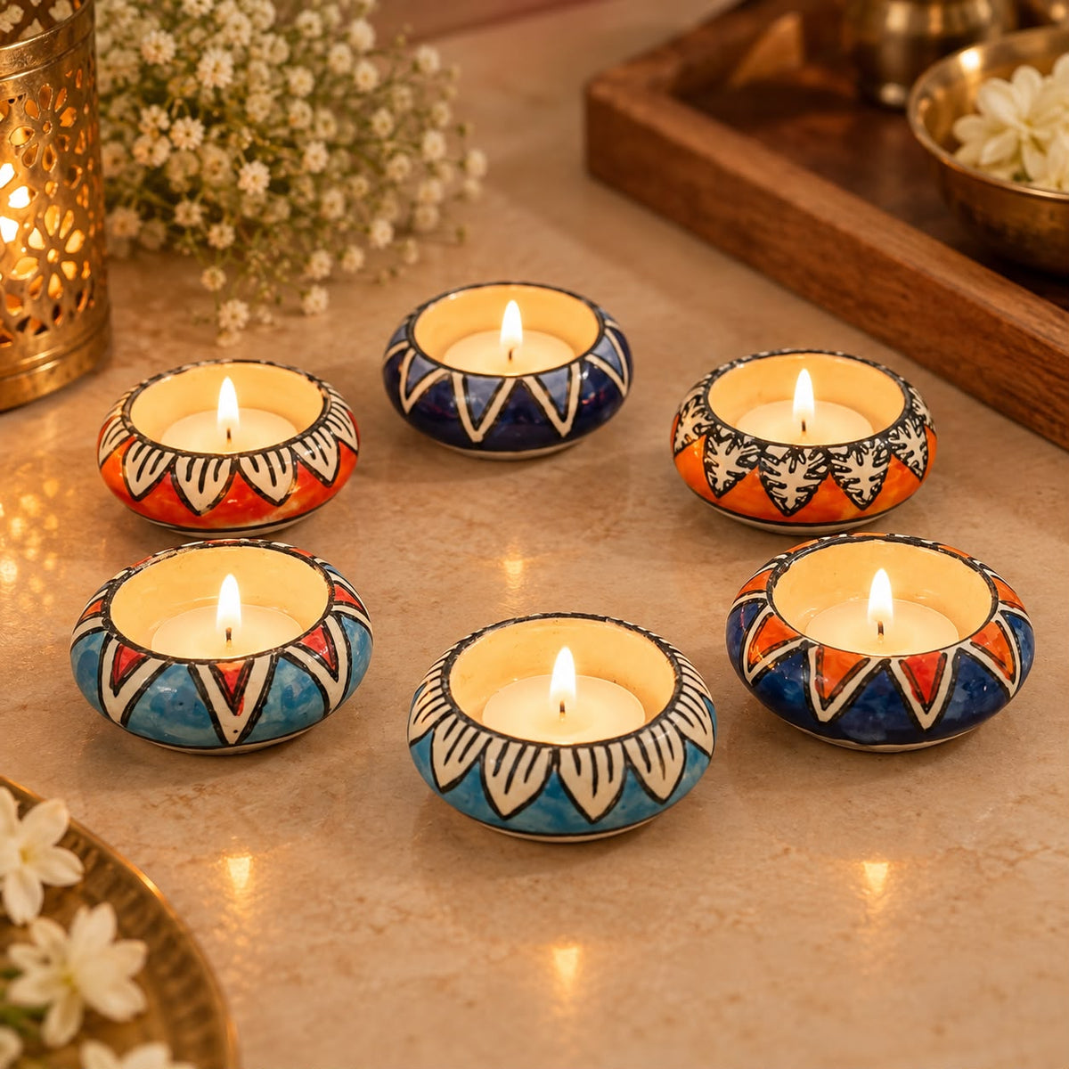 Six decorative tealight candles in hand-painted ceramic holders arranged on a beige stone surface. The holders feature colorful geometric patterns in blue, orange, red, and white, with the candles lit and glowing softly.
