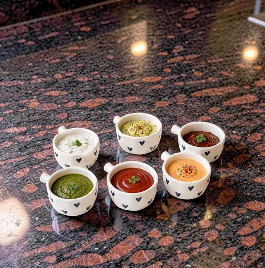 Six small white ceramic dipping cups with black heart patterns, filled with various dips like green chutney, white raita, pistachio chutney, tomato chutney, orange chutney, and a dark brown sauce, arranged on a dark speckled granite countertop.