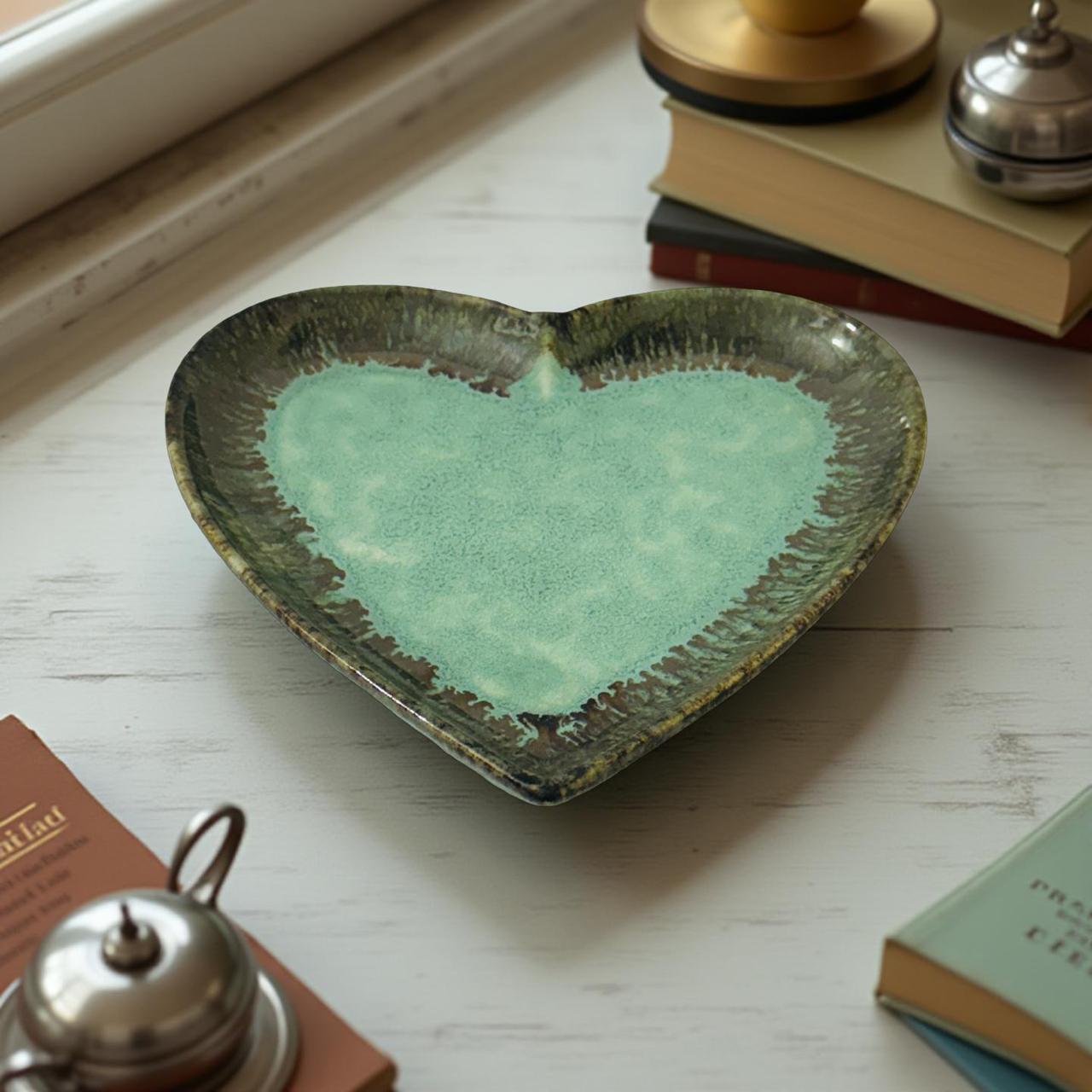 Heart-shaped ceramic dish with green glaze on a wooden surface with books and a teapot.