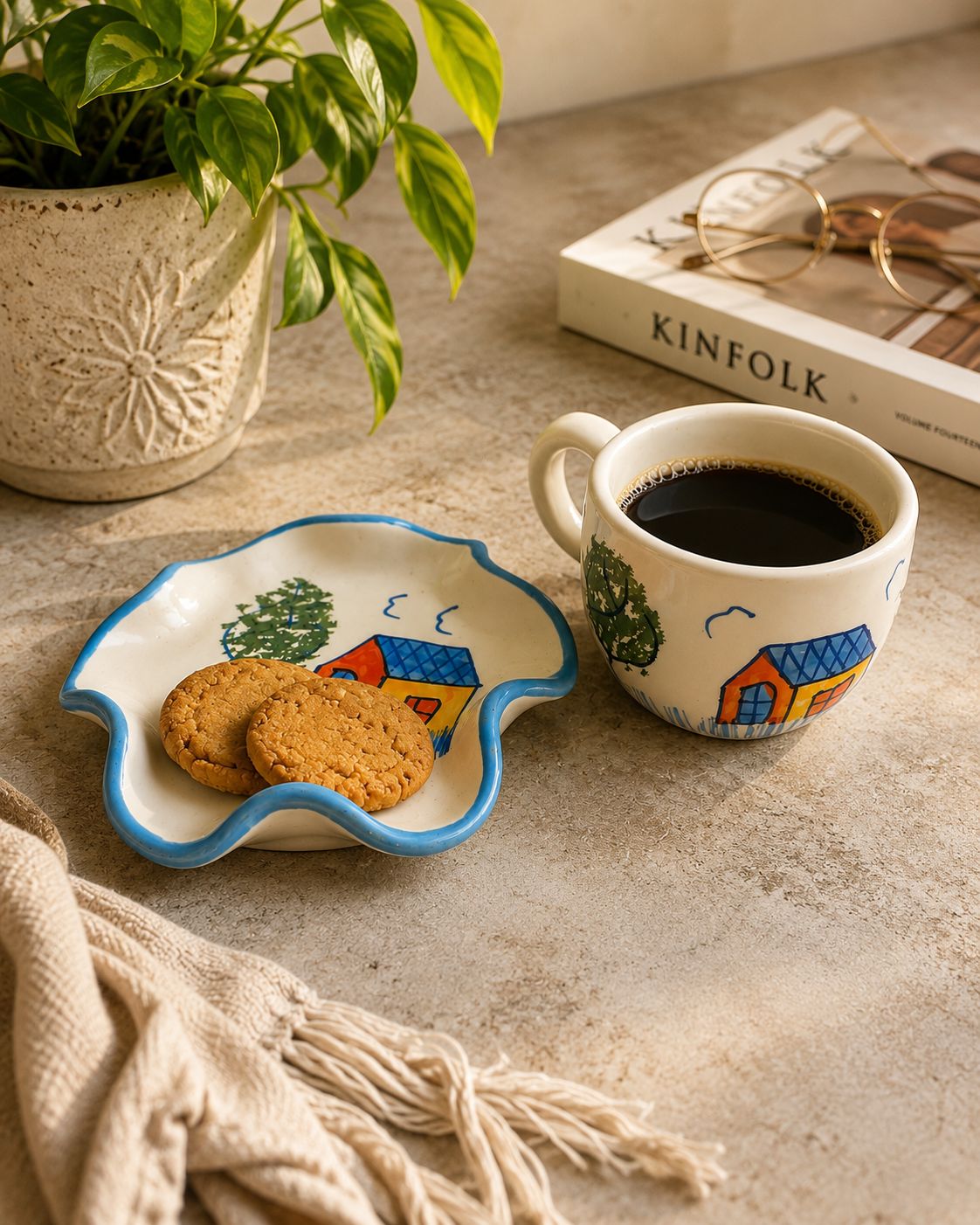 A styled tabletop scene with a small scalloped ceramic dish holding two cookies, a matching ceramic mug filled with black coffee, a potted green plant, a book, and a folded cream textile on a beige stone surface.