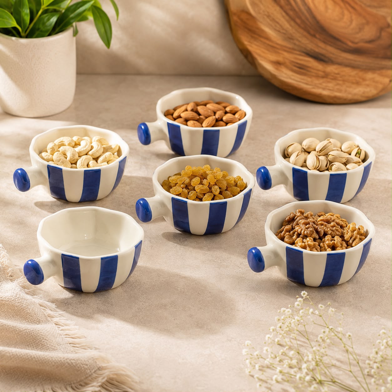 Six small white ceramic bowls with blue vertical stripes and blue handles are arranged on a beige fabric surface. Some bowls contain nuts and dried fruits, while one bowl is empty.