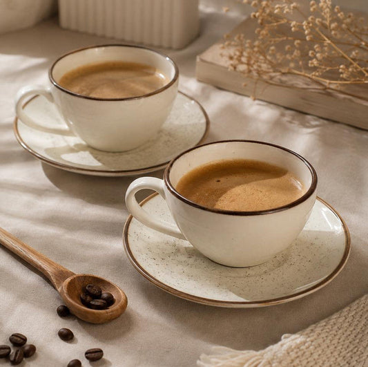 Two white ceramic coffee cups with saucers on a beige fabric surface, styled with coffee beans, a wooden spoon, and dried flowers.