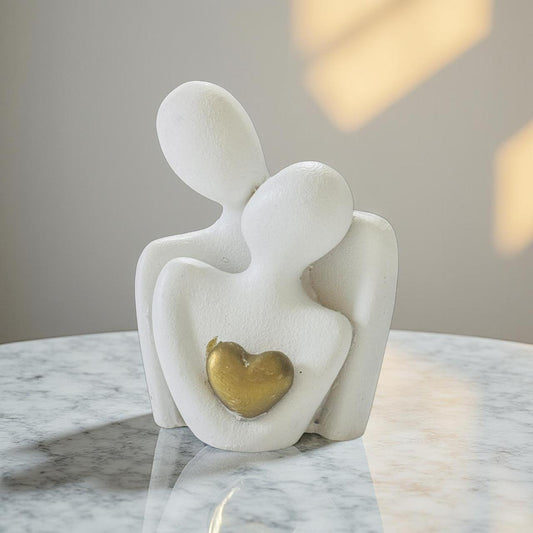 White abstract sculpture of two figures embracing with a gold heart on a marble surface.