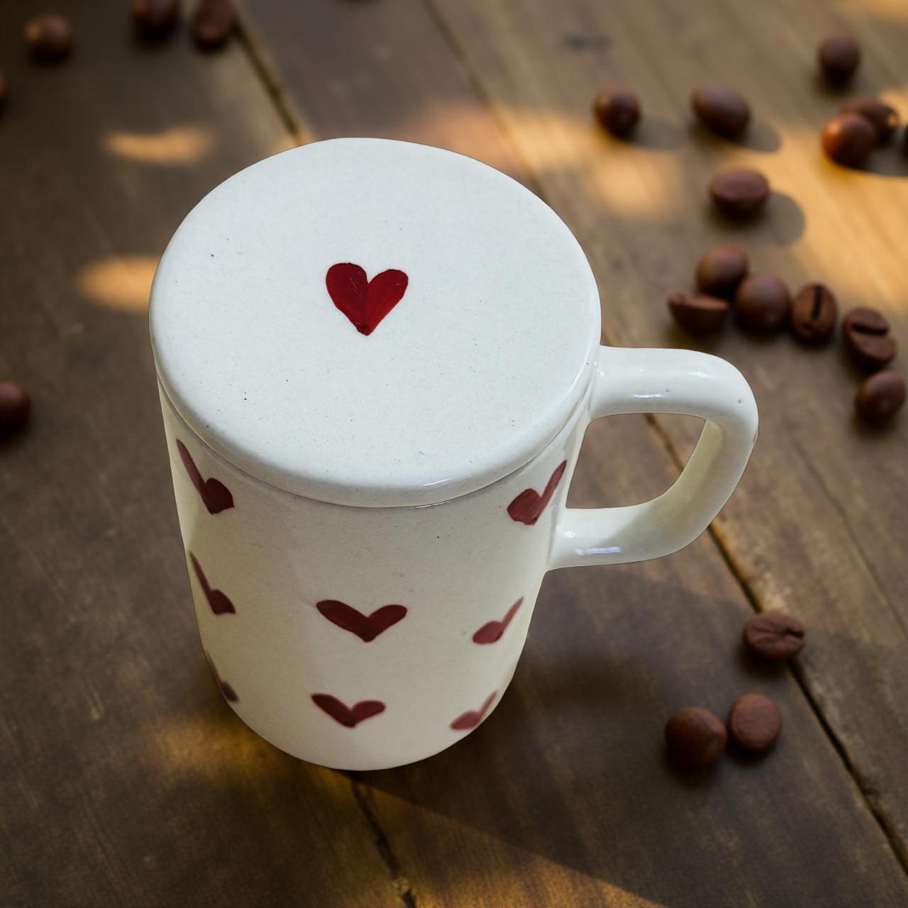 White mug with red heart design on lid and body, surrounded by coffee beans on a wooden surface