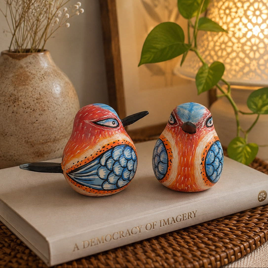 Two hand-painted wooden bird figurines with orange, blue, and white patterns, displayed on a book in a warm, styled home setting with a plant and lamp.