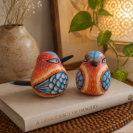 Two hand-painted wooden bird figurines with orange, blue, and white patterns, displayed on a book in a warm, styled home setting with a plant and lamp.
