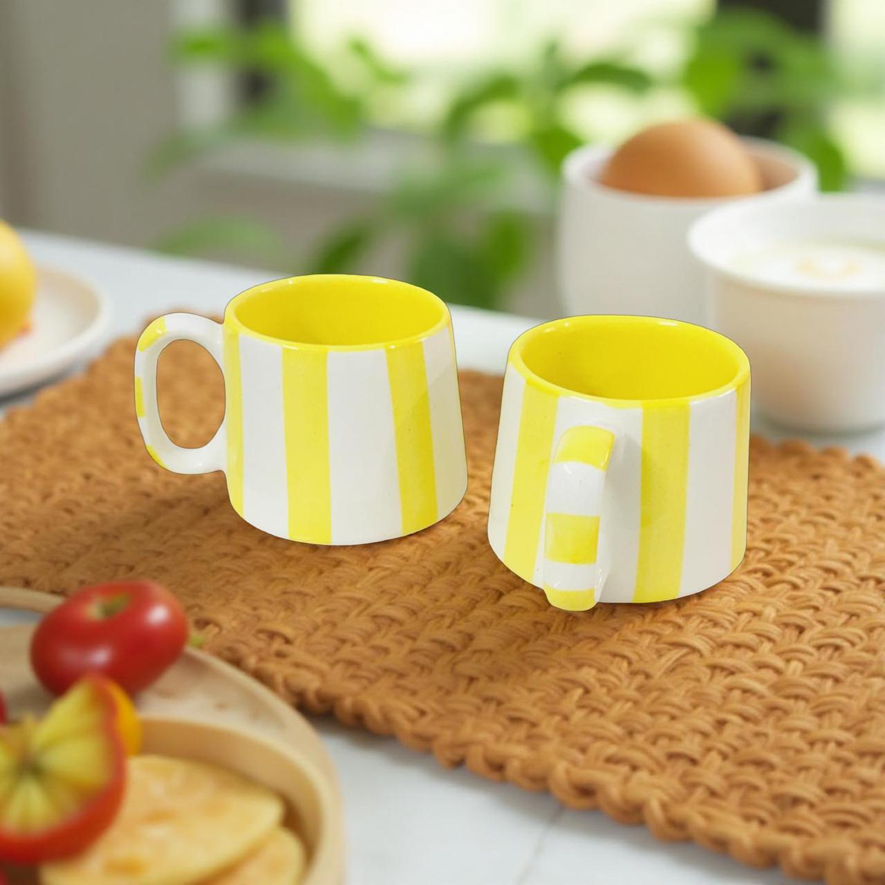 Two yellow and white striped mugs on a woven placemat with a blurred background of food items.